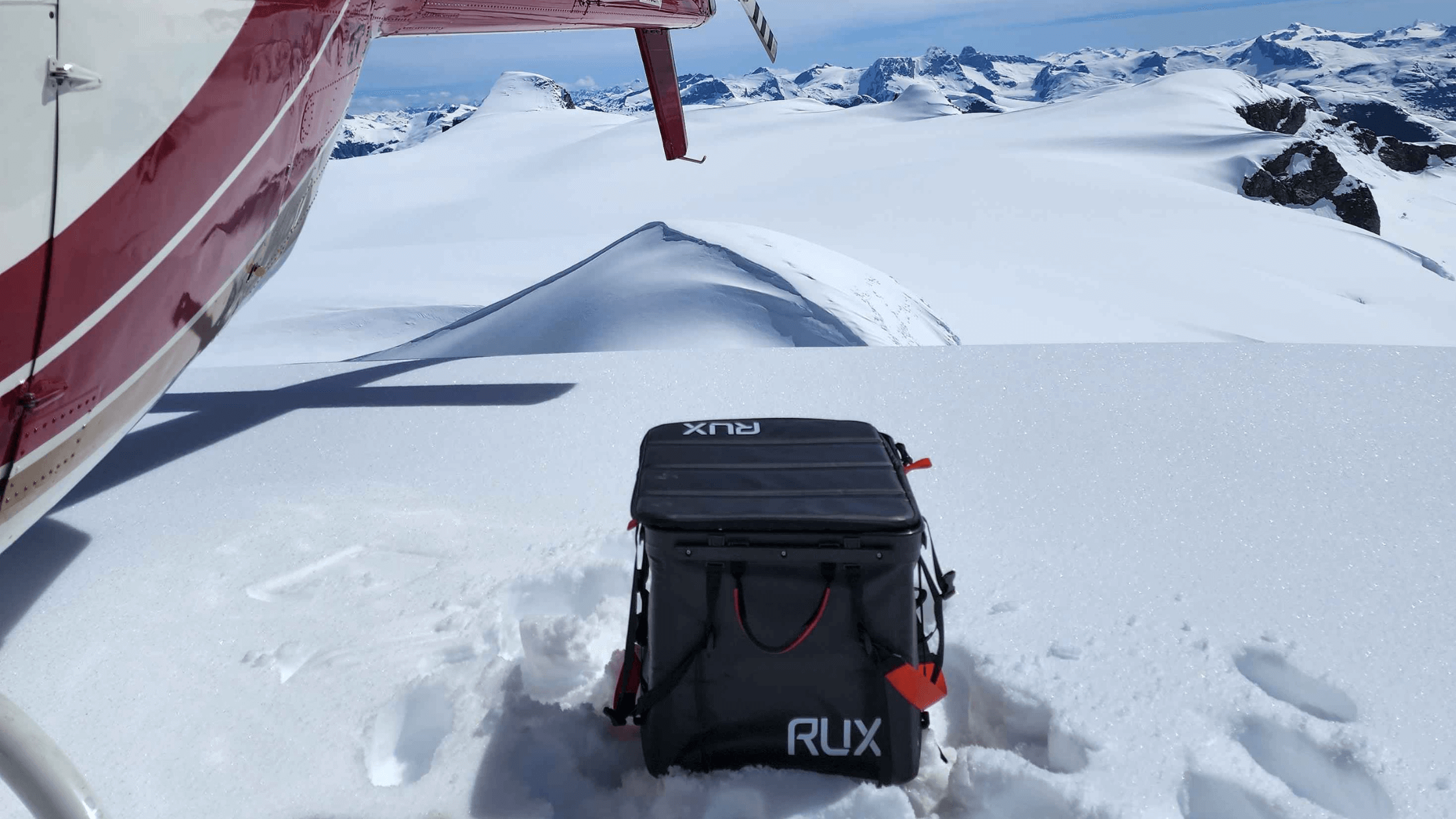 Top of snowy mountains, Black RUX 70L next to helicopter. 